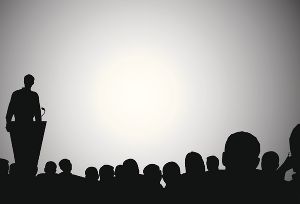 Abgebildet wird am linken Bildrand eine vortragende Person und am unteren Bildrand ein zuhörendes Publikum. Die Personen sind gänzlich schwarz. Der Hintergrund ist grau.